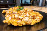 Almond Chicken Schnitzel Almond Chicken Schnitzel cutlet coated in golden sliced almonds, garnished with parsley on a black plate.