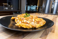 Golden Almond Chicken Schnitzel topped with parsley on a black plate over a wooden counter.