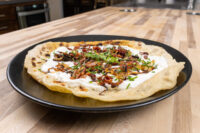 Lokše Flat Bread with Bryndza Cheese Substitute and Bacon Lokše Flat Bread with Bryndza Cheese Substitute and Bacon topped with fried onions and chives on a black plate.
