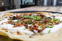 Lokše Flat Bread with Bryndza Cheese Substitute and Bacon Lokše Flat Bread with Bryndza Cheese Substitute and Bacon topped with onions, bacon bits, and chives on a dark board.