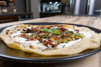 Lokše Flat Bread with Bryndza Cheese Substitute and Bacon Lokše Flat Bread with Bryndza Cheese Substitute and Bacon on a black plate, topped with cream, onions, and herbs.