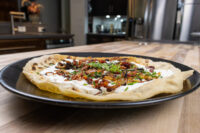 Lokše Flat Bread with Bryndza Cheese Substitute and Bacon Lokše Flat Bread with Bryndza Cheese Substitute and Bacon topped with cream, crispy bits, and chives on a black plate.
