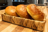 Burger Buns Three golden Burger Buns with sesame seeds sit side by side in a woven basket on a wooden counter.