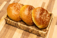 Burger Buns Three golden-brown Burger Buns topped with sesame seeds, arranged in a woven basket on a wooden table.