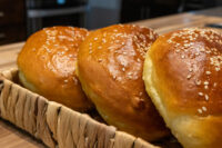 Golden Sesame Burger Buns Three golden Burger Buns topped with sesame seeds sit close together in a woven basket