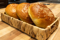 Burger Buns Three golden Burger Buns topped with sesame seeds sit in a woven basket on a wooden counter.
