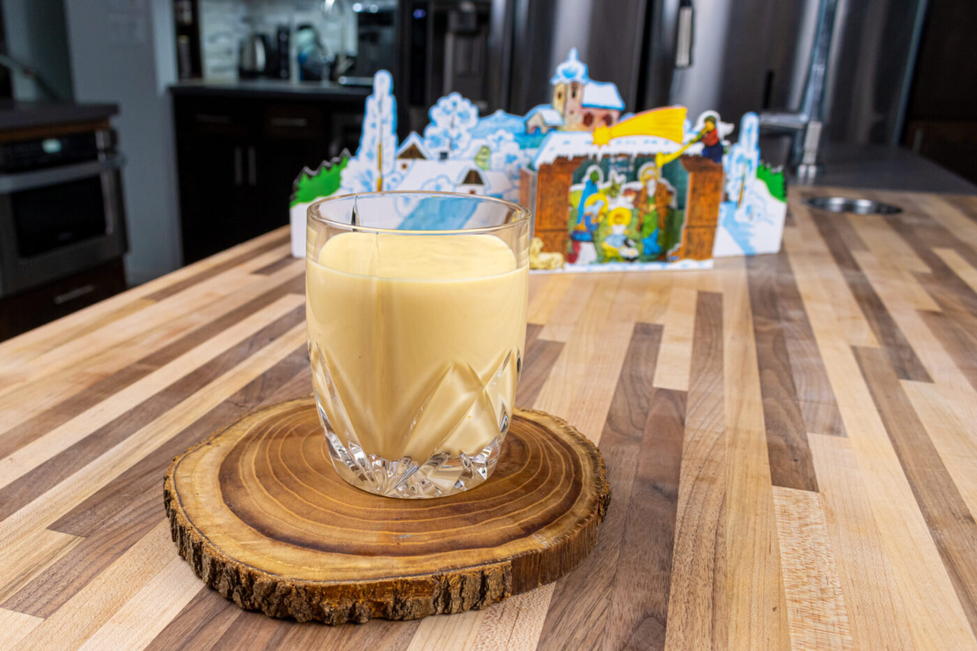 Czech Egg Liqueur – Czech Eggnog A glass of Czech Egg Liqueur – Czech Eggnog on a wooden coaster with a winter village decoration behind it.