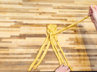 Hands braiding strands of yellow dough into Christmas Vánočka (Houska) on a wooden board.