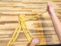 Hands braiding strands of yellow dough at a knotted center for Christmas Vánočka (Houska).
