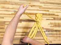 Hands braiding strands of yellow dough into Christmas Vánočka (Houska) on a wooden board.