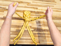 Hands shaping raisin-studded dough strands into a braid for Christmas Vánočka (Houska) on a wood board.