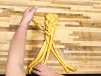 Hands braiding strands of yellow dough into a partial braid for Christmas Vánočka (Houska).