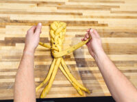 Hands braiding strands of raisin-studded dough into a Christmas Vánočka (Houska) on a wooden board.
