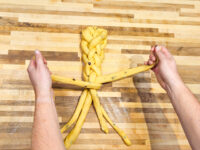 Hands braiding strands of raisin-studded dough into a Christmas Vánočka (Houska) loaf on a wooden board.