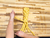 Hands braiding strands of yellow dough with raisins into a Christmas Vánočka (Houska) on a wooden board