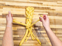 Hands braid strands of yellow dough into a Christmas Vánočka (Houska) on a wooden board.