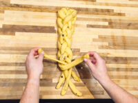 Hands braiding strands of dough for Christmas Vánočka (Houska) on a wooden board.