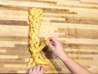 Hands braid strands of dough into a loaf for Christmas Vánočka (Houska) on a wooden board.