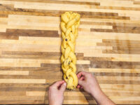 Hands braiding a strand of yellow Vánočka (Houska) dough dotted with raisins on a wooden board.
