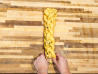 Hands braiding raisin-studded dough into a strand for Christmas Vánočka (Houska) on a wooden board.