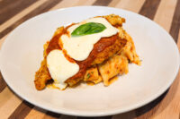 Chicken Parmesan Chicken Parmesan cutlet topped with marinara, melted mozzarella, and a basil leaf, served over wide pasta ribbons on a white plate