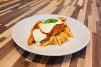Chicken Parmesan Chicken Parmesan cutlet topped with marinara and melted mozzarella, garnished with basil, served over pasta on a white plate.
