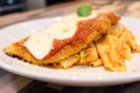 Chicken Parmesan Breaded Chicken Parmesan topped with marinara and melted cheese, served beside ribbon pasta on a white plate.