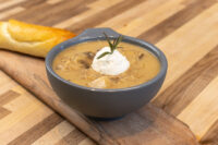 Gray bowl of Valašská Kyselica Soup topped with sour cream and rosemary, bread on the side.