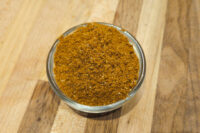 Small glass bowl filled with Garam Masala Seasoning, a reddish-brown ground spice blend, on a wooden surface.