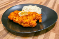 Cheese Battered Chicken Schnitzel Kuře v Sýrovem Těstíčku with a lemon wedge and coleslaw on a black plate