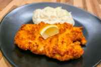 Golden-brown Cheese Battered Chicken Schnitzel Kuře v Sýrovem Těstíčku plated with a lemon wedge and a scoop of mashed potatoes on a dark blue dish.