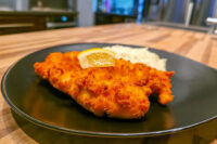 Cheese Battered Chicken Schnitzel Kuře v Sýrovem Těstíčku with lemon wedge and rice on a black plate