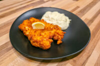 Golden-brown Cheese Battered Chicken Schnitzel Kuře v Sýrovem Těstíčku on a black plate with mashed potatoes and a lemon wedge.
