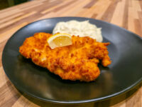 Golden breaded Chicken Schnitzel Kuře v Sýrovém Těstíčku with a lemon wedge and creamy side on a dark plate.