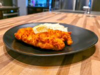 Golden fried Cheese Battered Chicken Schnitzel Kuře v Sýrovem Těstíčku with a lemon wedge and mashed potato on a black plate.