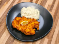 Golden fried Cheese Battered Chicken Schnitzel Kuře v Sýrovem Těstíčku with lemon wedge and mashed potato on a dark plate.