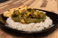 Spinach Palak Paneer Black plate of Spinach Palak Paneer over white rice with grilled naan bread on the side.