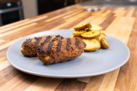 Two grilled Čevapčiči Sausages with char marks on a grey plate beside roasted potato wedges