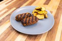 Two grilled Čevapčiči Sausages With Cheese with char lines beside roasted potato slices on a grey plate