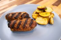 Two grilled Čevapčiči Sausages With Cheese with char marks beside roasted potato wedges on a grey plate