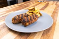 Two grilled Čevapčiči Sausages With Cheese with char marks, served with roasted potato wedges on a gray plate.