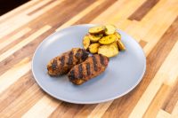 Two grilled Čevapčiči sausages with char marks on a gray plate beside roasted potato slices