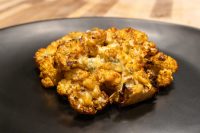 Roasted Buffalo Cauliflower Steak florets glazed orange-red with melted cheese, charred edges, on black plate