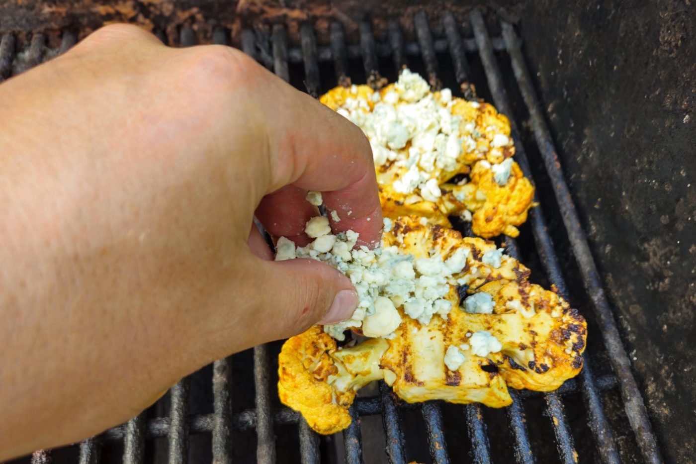 Hand sprinkles crumbled blue cheese over charred Buffalo Cauliflower Steak on a grill grate.