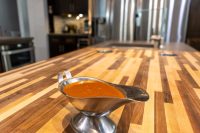 Buffalo Sauce Recipe A metal gravy boat filled with glossy orange-red Buffalo Sauce sits on a striped wooden countertop.