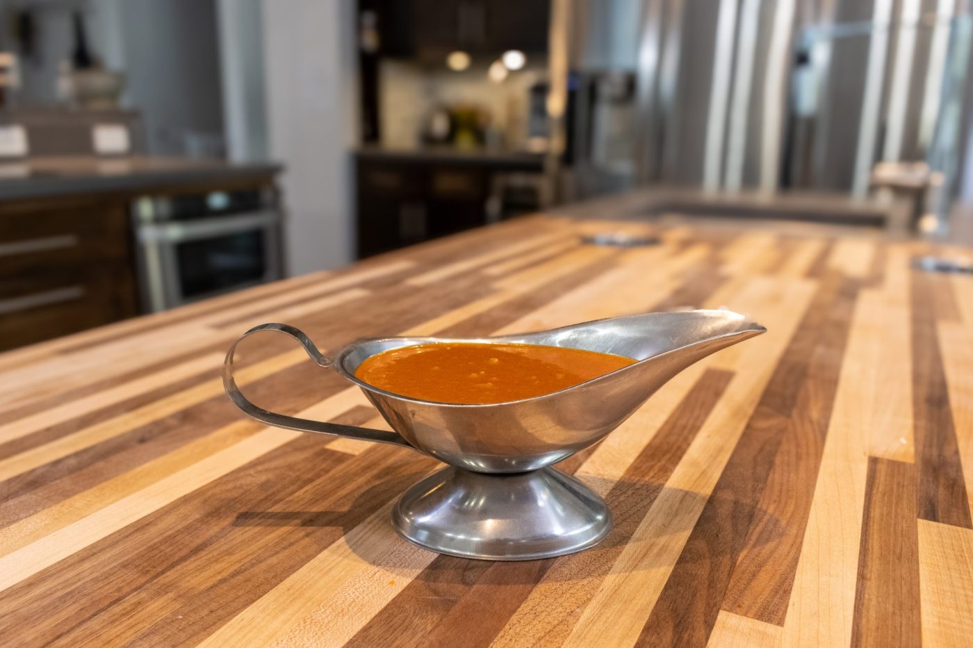 Buffalo Sauce Recipe Orange-red Buffalo Sauce served in a metal gravy boat on a striped wooden countertop.