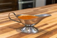 Buffalo Sauce in Gravy Boat Buffalo Sauce pooled in a stainless steel gravy boat on a wooden countertop.