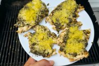 Pesto Chicken Breasts Plated Four Pesto Chicken Breasts topped with green pesto and yellow parmesan on a white plate near a grill.