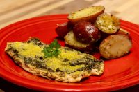 Pesto Chicken Breasts Pesto Chicken Breasts coated in green pesto and parmesan, garnished with parsley, on a red plate with potatoes.