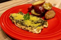 Pesto Chicken Breasts Pesto Chicken Breasts topped with grated parmesan and parsley, served with roasted red potatoes on a red plate.